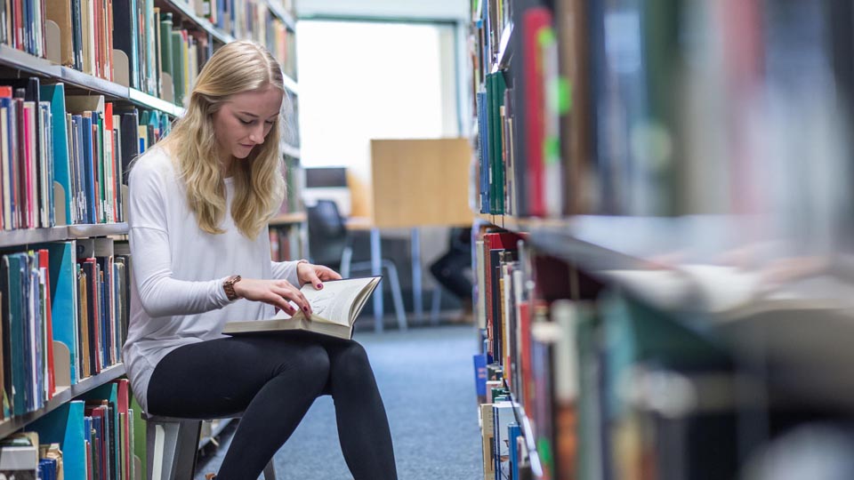 a student sitting reading next to shelves of books in the library